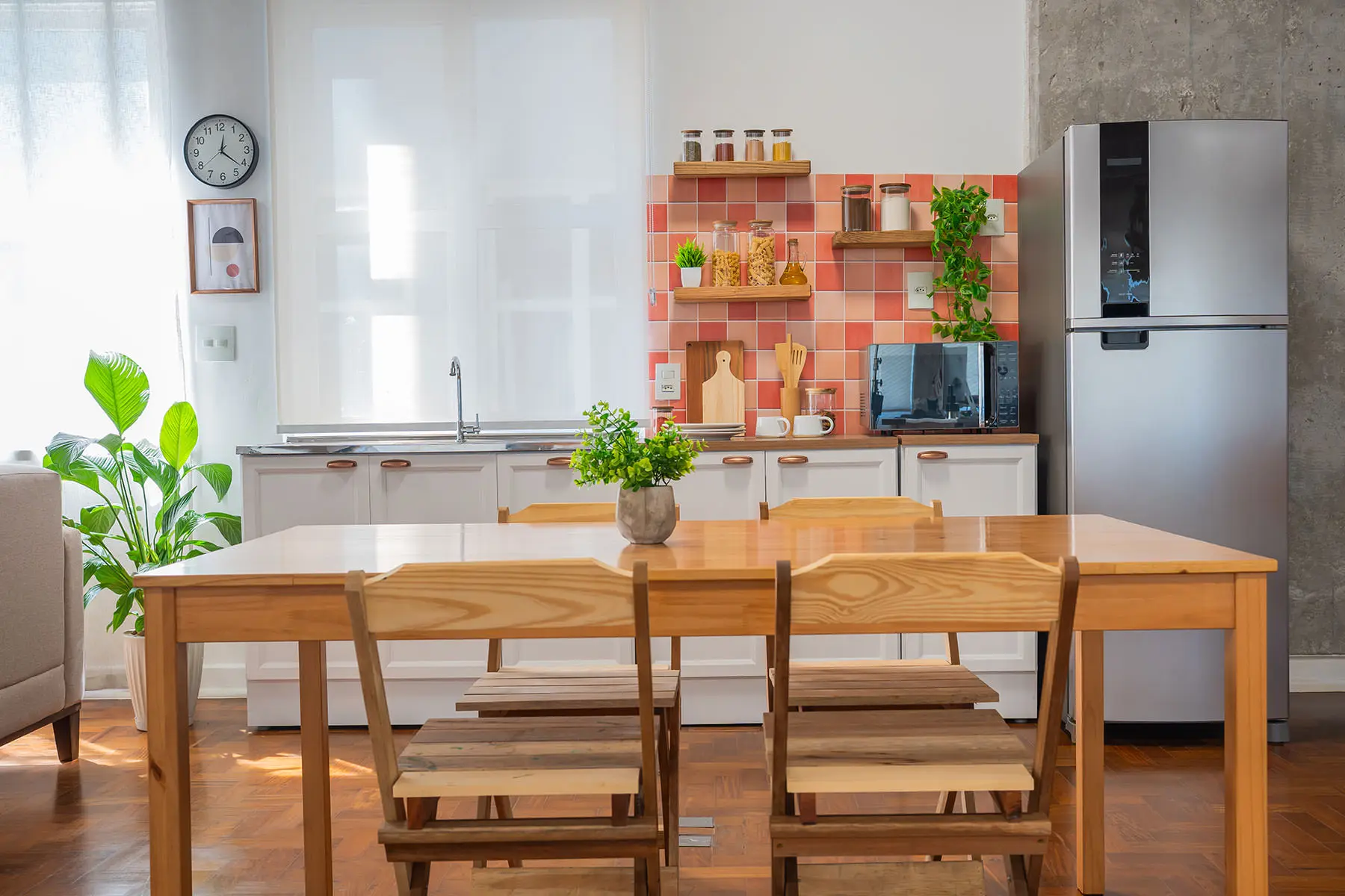 Ambiente cozinha do estúdio: bancada branca livre, armários, geladeira, painel de azulejo laranja e mesa de madeira com cadeiras